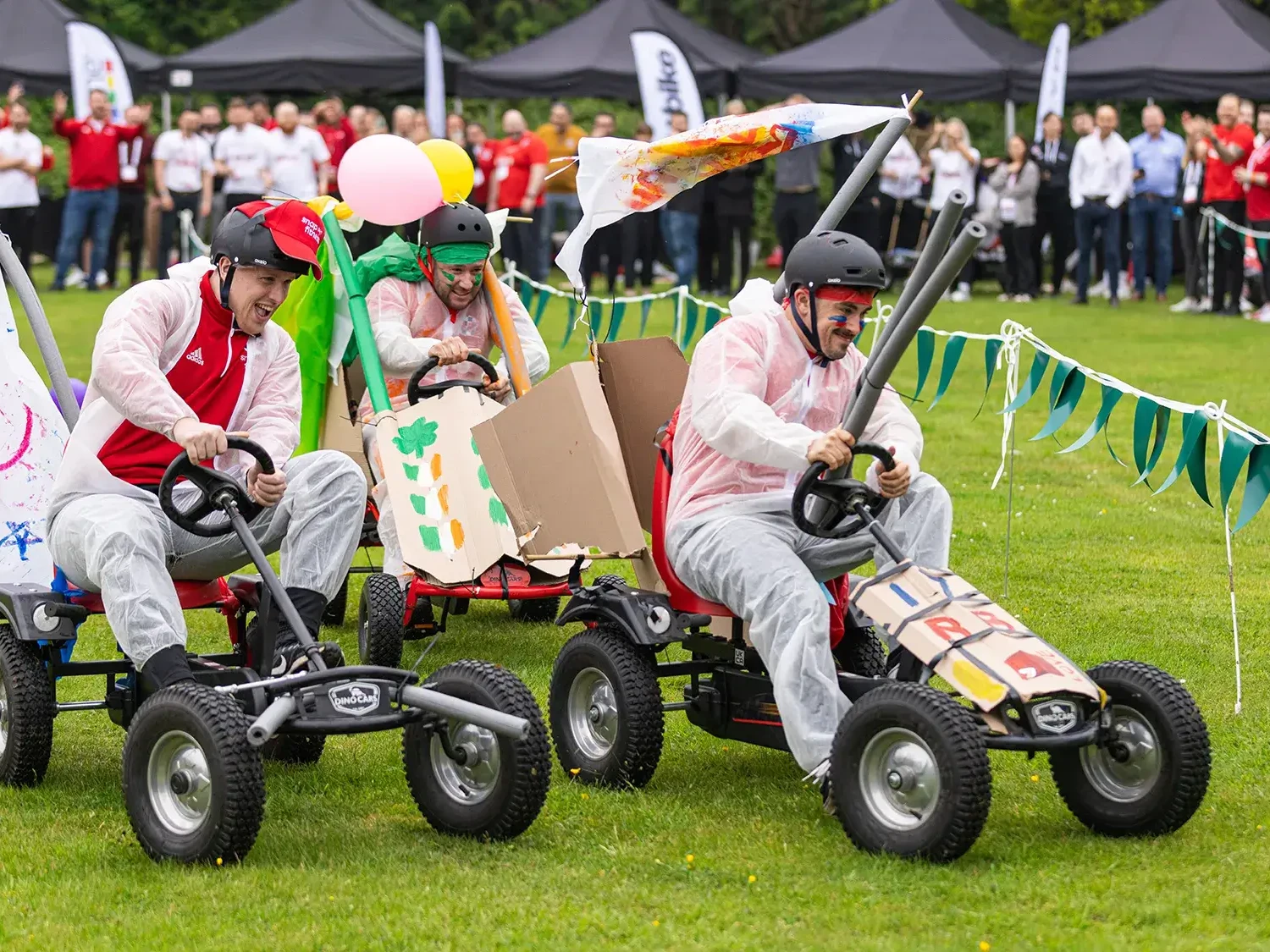 Team building participants racing fun carts on grass surrounded by spectators