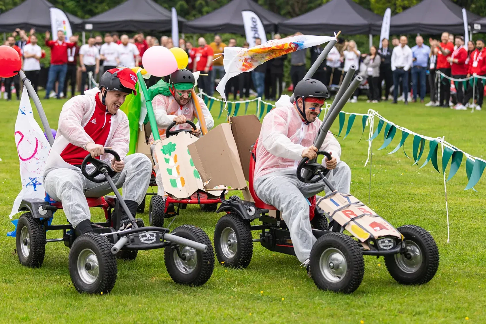 Team building participants racing fun carts on grass surrounded by spectators