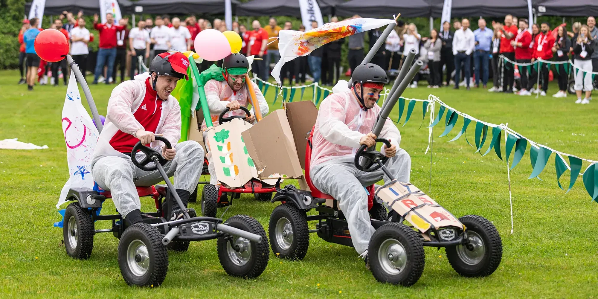 Team building participants racing fun carts on grass surrounded by spectators